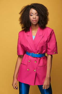Beauty portrait of attractive young african girl with afro hairstyle. Girl wearing pink jacket, blue leggings and belt posing on yellow background. Studio shot