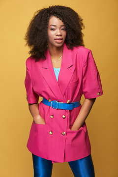 Beauty portrait of attractive young african girl with afro hairstyle. Amazing girl wearing pink jacket, blue leggings and belt posing on yellow background. Studio shot.