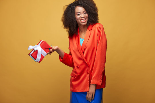 Bright portrait of young african girl with afro hairstyle. Smiling girl wearing orange jacket, dark blue latex skirt holds red present in her hand and posing on yellow background. Studio shot.