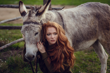 A girl with curly red hair in fashionable clothes in the style of Provence hugs a cute donkey