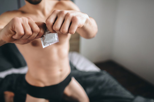 Safety sex concept. Cropped image of a naked bearded dark-hair handsome smiling man holding condom in hand on bed