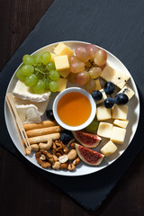 plate of cheeses, snacks and fruits on a dark background, vertical top view