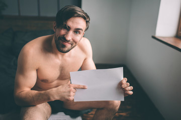 Naked bearded dark-hair handsome manholding an empty sheet of paper on the bed. Home Interier
