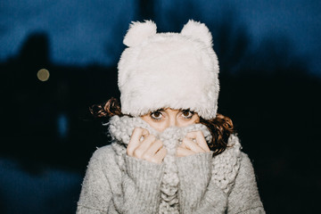 .Portrait of a young and pretty woman in the middle of winter night. Sheltered with hat and scarf, getting very cold. Lifestyle..