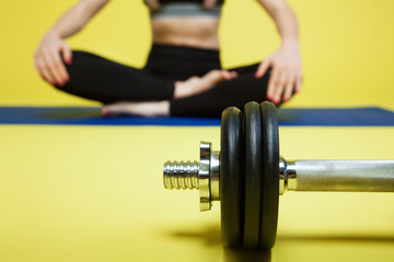 Fototapeta na wymiar Young sports woman sits on the floor in pose of lotus near the dumbbells, at the gym. yellow background