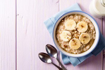 Oatmeal porridge with banana, walnuts and honey in bowl on purple wooden background. Healthy breakfast.