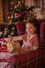 Fototapeta na wymiar cute girl sitting in a chair by christmas tree opening christmas present gift 