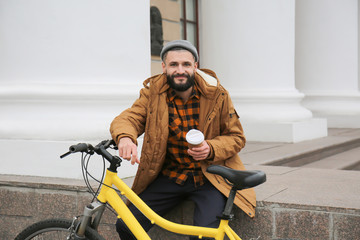 Attractive young hipster with cup of coffee and bicycle on city street