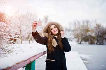 Beautiful brunette girl in winter warm clothing. Model on winter jacket against frozen lake at park making selfie on phone.