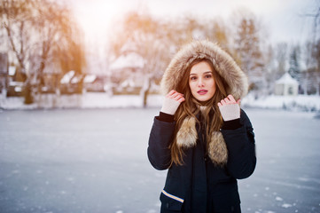 Beautiful brunette girl in winter warm clothing. Model on winter jacket against frozen lake at park.