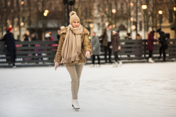 Naklejka premium Full length portrait of young female with blonde hair in fur coat, beige hat, scarf and trousers skating on ice rink, outdoors at winter / Weekends activities outdoor in cold weather/