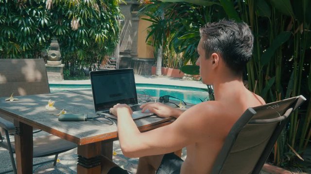 young male working on laptop. The guy freelancer works in a beautiful place in the resort near the pool