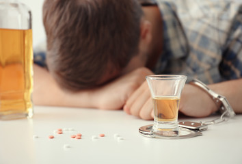 Drunk man in handcuffs sitting at table with bottle and glass of brandy. Alcohol dependence concept