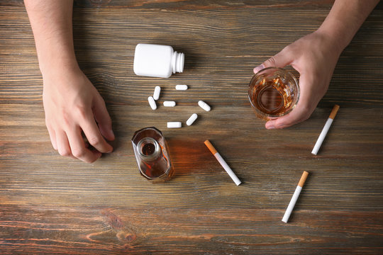 Man sitting at table with cigarettes, drugs and alcohol at wooden table. Concept of bad habits
