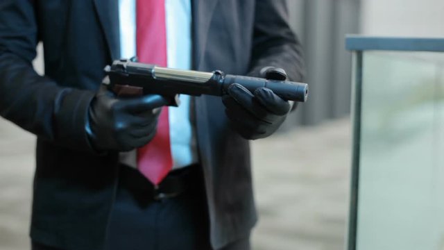 Hands in black gloves screw the muffler onto the gun, it's a hired killer.