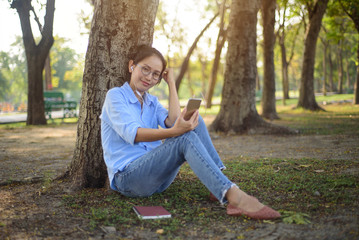 Naklejka na ściany i meble Asian woman listening to music from smartphone under the tree in the garden.