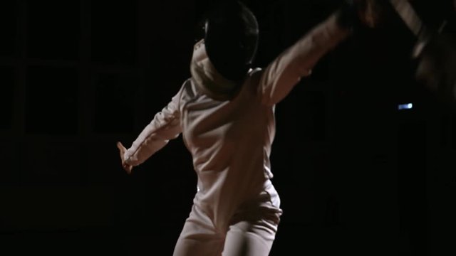 Young women and men in hats and suits for fencing, fighting with swords. Dark background. The camera moves behind the men