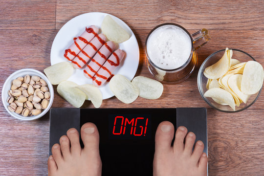 Male feet on digital scales with word omg on screen. Beer and plates with junk food (sausages, potato chips, pistachios). Concept of unhealthy eating and alcohol drinking.