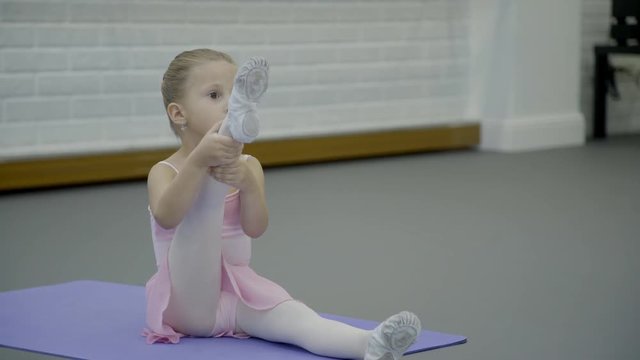 Small girl performs ballet movement in dance studio indoors. Little princess is at choreographic class in ballet school. Beginning ballerina in silk costume practices intensely, sitting on gymnastic