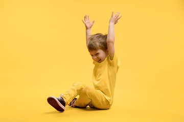 Happy boy in yellow clothes sitting and rising hands up at camera against yellow background