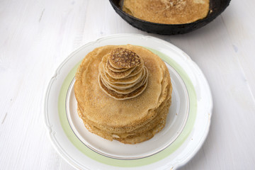 Large and small pancakes are located on a light wooden table. Frying pan in which pancakes were prepared on the back of a blurry background.