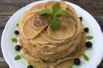 Large and small pancakes are located in a plate on an old wooden table decorated with berries and mint leaves. Close-up.