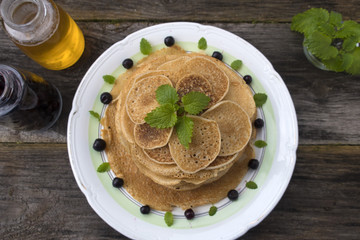 Large and small pancakes are located in a plate on an old wooden table decorated with berries and mint leaves.