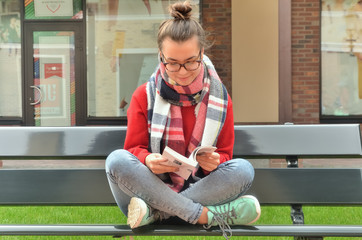 Asian girl in glasses sits on a bench, crosses her legs and reads a book