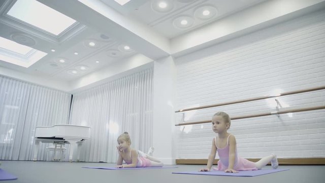 Two girls do arch laying on the mats while ballet class with famous trainer. Children stretch their heads to their feets in this position very diligently. Little dancers are taught in big bright