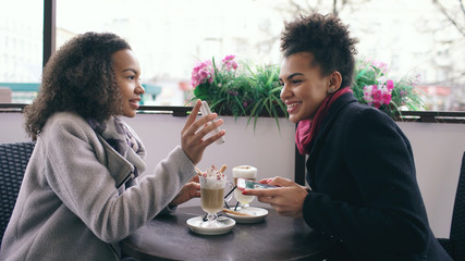 Two attractive mixed race female friends sharing together using smartphone in street cafe outdoors