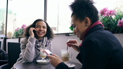 Two mixed race women sitting at the table in the street cafe using smartphone and talking