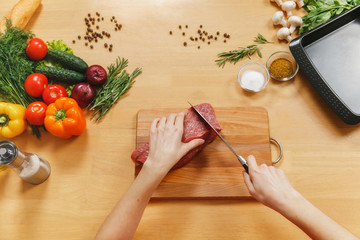 Raw meat. Fresh beef tenderloin on cutting board on wooden table with different vegetables, spices, mushrooms, baking tray, knife. Top view flat lay Copy space for advertisement. Woman cuts with knife