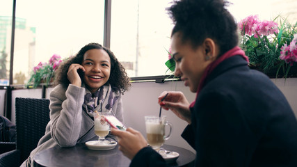 Two mixed race women sitting at the table in the street cafe using smartphone and talking
