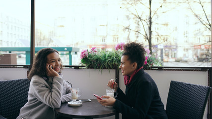 Attrcative mixed race woman sitting at the table in the street cafe talking cell phone while her friend surfing smartphone