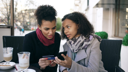 Two attractive mixed race women having online shopping with credit card and smartphone while talking and drinking coffee in street cafe
