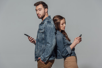 young couple in denim jackets standing back to back and using smartphones isolated on grey