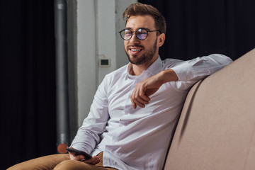 handsome smiling young man in eyeglasses holding smartphone and looking away at home