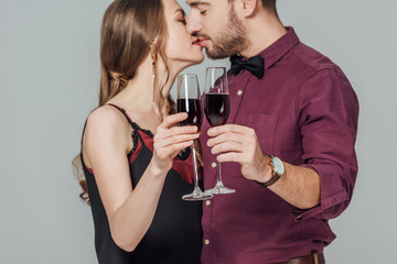 close-up view of beautiful young couple holding glasses of red wine and kissing isolated on grey