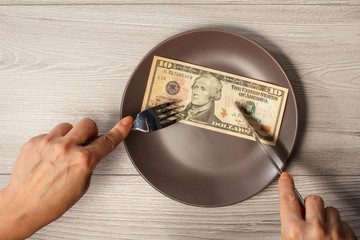 Ten American dollars bill on brawn plate and male hands with fork and knife. Money ready to be eaten