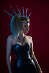 Fototapeta premium Blonde woman in black leather corset, skirt and hat with thorns posing against red background