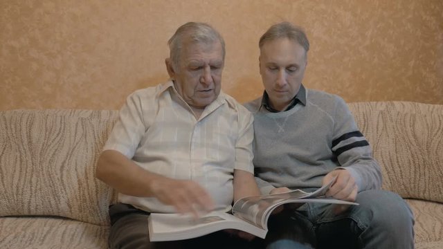 An elderly man is looking at a book with his grown-up son.