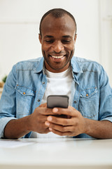 Communicating. Attractive alert dark-haired afro-american man smiling and writing a message on his phone while sitting at the table