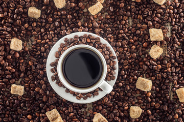top view of cup of coffee with saucer, roasted coffee beans and brown sugar on sackcloth