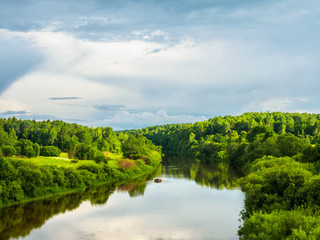 Fototapeta premium Bend Of Riverbed Of Small River With Thick Green Forest Growing On Its Bank Illuminated By Evening Sun