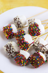 marshmallows on a sticks, laying on a white plate. glazed with chocolate and colorful sprinkles.