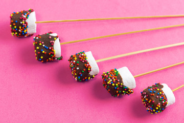 marshmallows on a sticks, laying on pink background. glazed with chocolate and colorful sprinkles.
