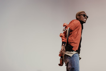 Stylish young african american skater holding longboard behind his back isolated on light background © LIGHTFIELD STUDIOS