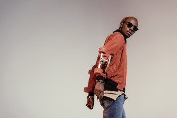 Stylish young african american skateboarder holding longboard behind his back isolated on light background