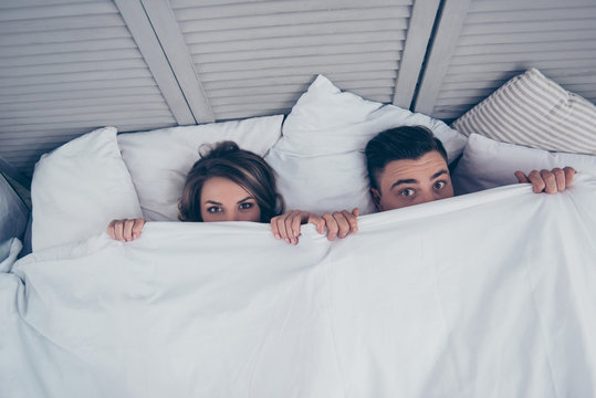 Top view of beautiful funny partners hiding behind blanket, peeking over sheets and looking at camera while lying in bed at home, weekend, rest, relax, someone catchees them