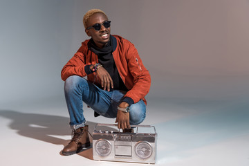 cheerful stylish young african american man crouching with silver tape recorder on grey